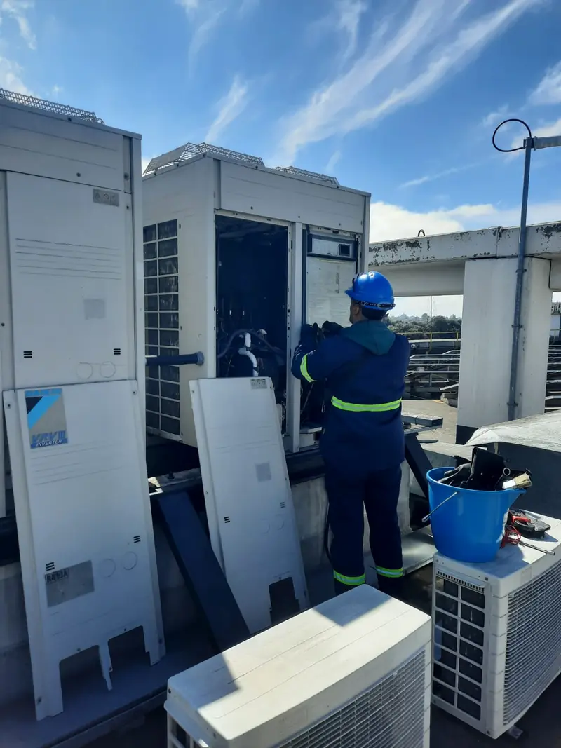 Técnico con casco y ropa de alta visibilidad trabajando sobre una unidad exterior VRV abierta en una terraza, junto a otras unidades del sistema